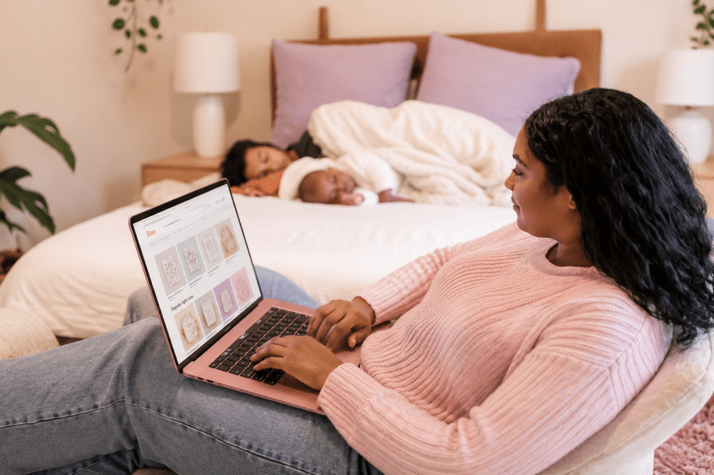 Mom browsing Etsy on a pink laptop while relaxing in a bedroom as her family sleeps nearby, illustrating the flexibility of building an online business from home.